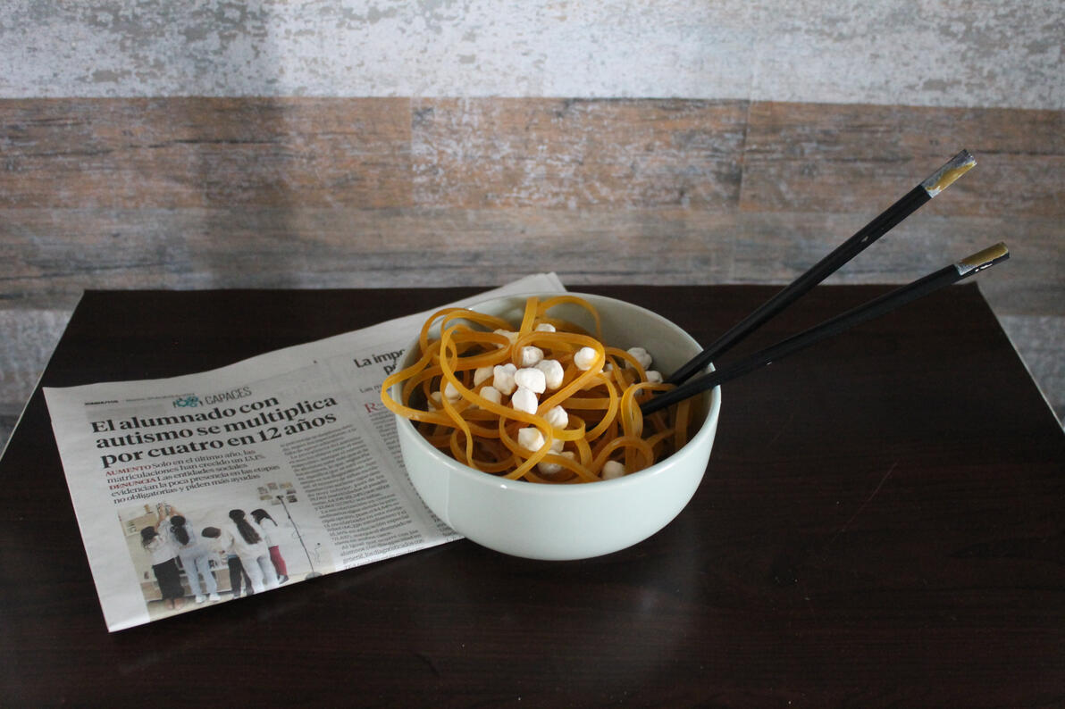 Ready made construido con objetos cotidianos; simula bol con spaguettis y garbanzso y palillos chinos encima de una revista cuyo titular es: El alumnado con autismo se multiplica por 4 en 12 años.